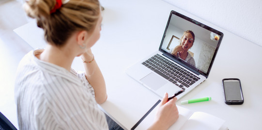 Unterstützung Am Schreibtisch sitzende Studentin schaut ein Lernvideo, daneben liegen Bücher und Stifte.